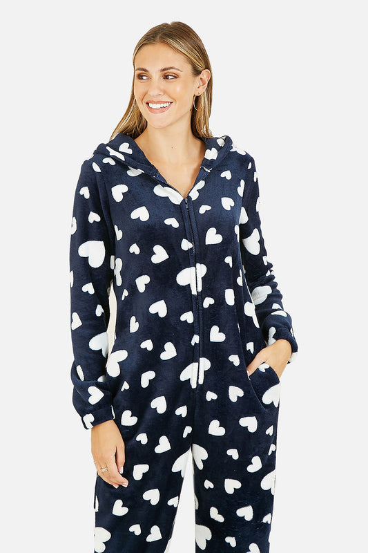 A woman wearing a navy blue hooded one-piece pajama with a white heart pattern, standing against a light gray studio background.